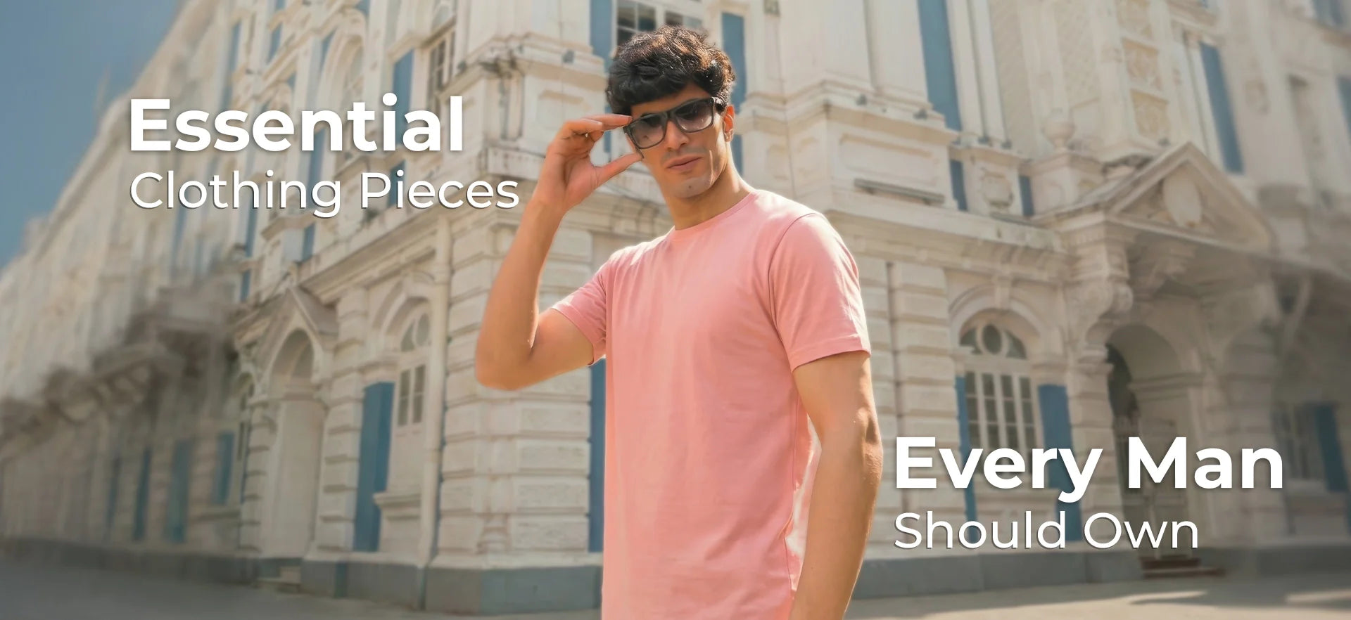Essential clothing for men: model in a pink t-shirt and sunglasses posing in front of a classic stone building.